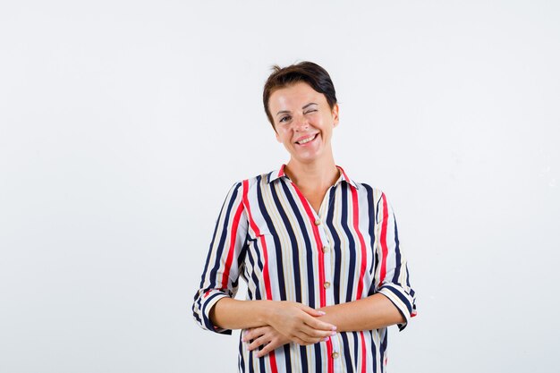 Mature woman posing with clasped hands, smiling, winking in striped blouse and looking sanguine. front view.