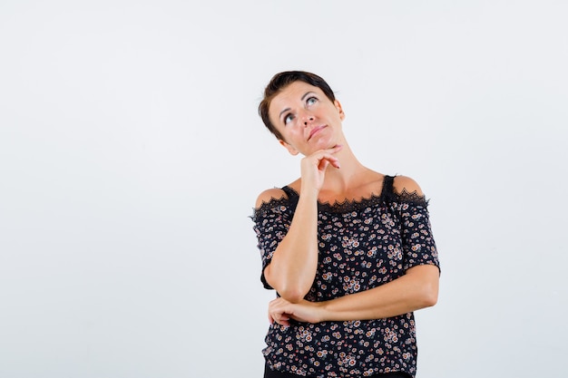 Mature woman propping chin on hand, holding hand under elbow, thinking about something in floral blouse, black skirt and looking pensive , front view.