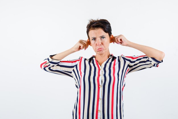 Mature woman pulling ears , curving lips in striped blouse and looking morose , front view.