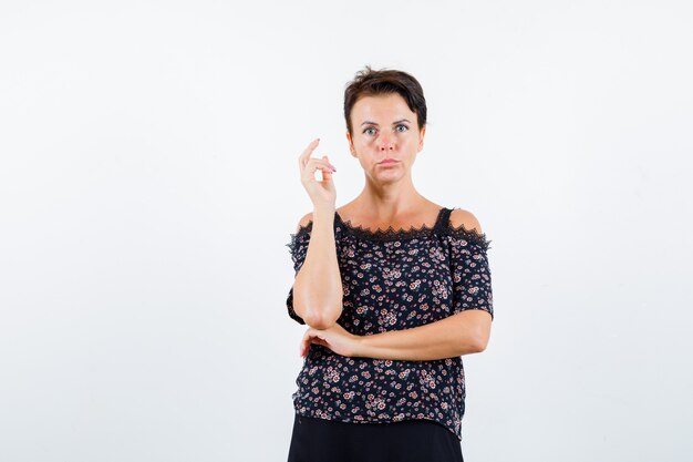 Mature woman raising hand, holding one hand under elbow in floral blouse and black skirt and looking serious. front view.