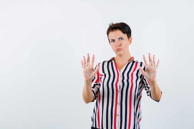 Mature woman raising palms in surrender gesture in striped blouse and looking scared. front view.