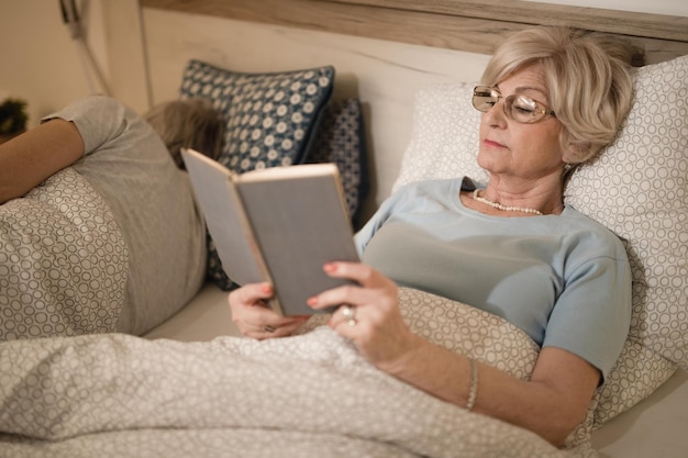 Mature woman reading book while lying down in bed in the evening Her husband is sleeping next to her