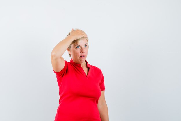 Mature woman in red t-shirt holding hand on head and looking confused.