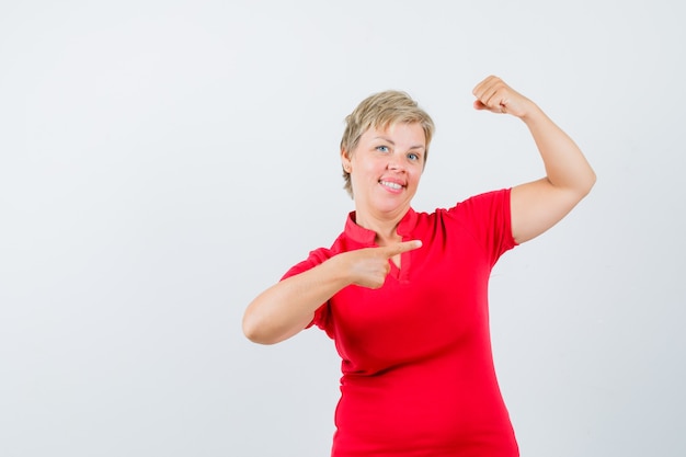 Mature woman in red t-shirt pointing at muscles of arm and looking confident.