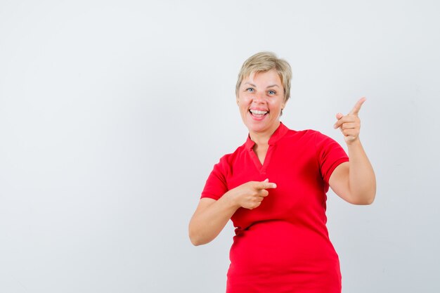 Зрелая женщина в красной футболке указывая на верхний правый угол и выглядит веселой.