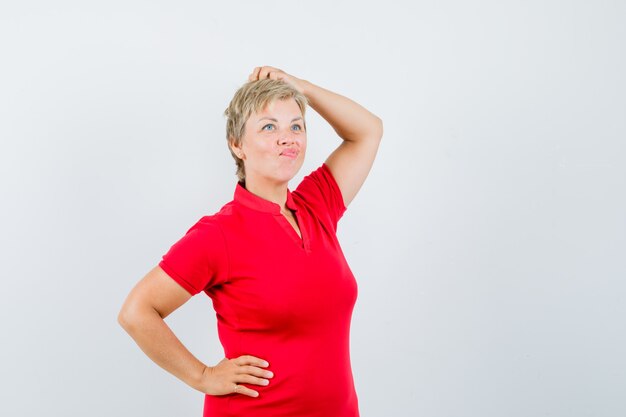 Mature woman in red t-shirt posing while scratching head and looking pensive.