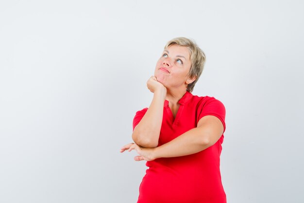 Зрелая женщина в красной футболке подпирает подбородок кулаком и смотрит задумчиво.