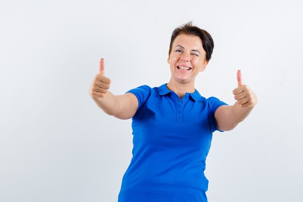 Mature woman showing double thumbs up in blue t-shirt and looking happy. front view.