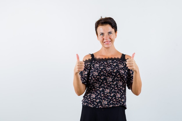 Mature woman showing double thumbs up in floral blouse and black skirt and looking cheerful , front view.