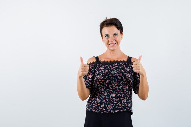 Mature woman showing double thumbs up in floral blouse and black skirt and looking cheerful , front view.