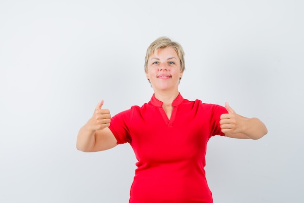 Зрелая женщина показывает двойные пальцы вверх в красной футболке и выглядит уверенно.