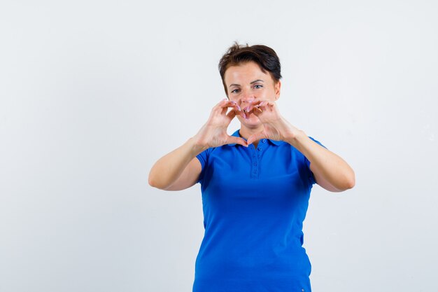 Mature woman showing heart gesture in blue t-shirt and looking merry , front view.