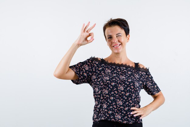 Mature woman showing ok sign, holding hand on waist, winking in floral blouse, black skirt and looking cheery , front view.