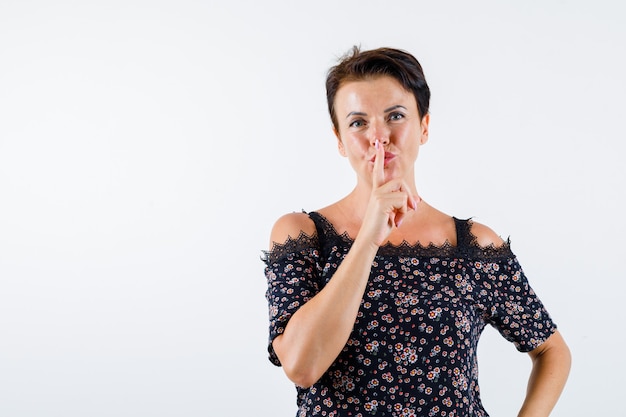 Mature woman showing silence gesture in floral blouse, black skirt and looking cheery , front view.