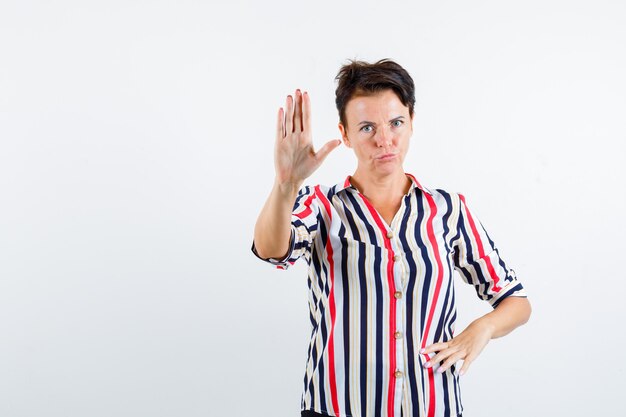 Mature woman showing stop sign, holding hand on waist in striped blouse and looking serious , front view.