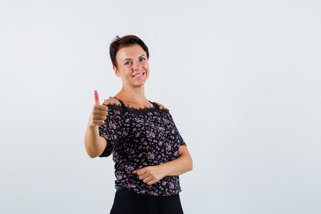 Mature woman showing thumb up in floral blouse and black skirt and looking cheerful , front view.