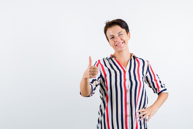 Mature woman showing thumb up, winking, holding hand on waist in striped blouse and looking confident. front view.