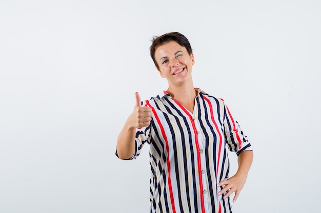 Mature woman showing thumb up, winking, holding hand on waist in striped blouse and looking confident. front view.
