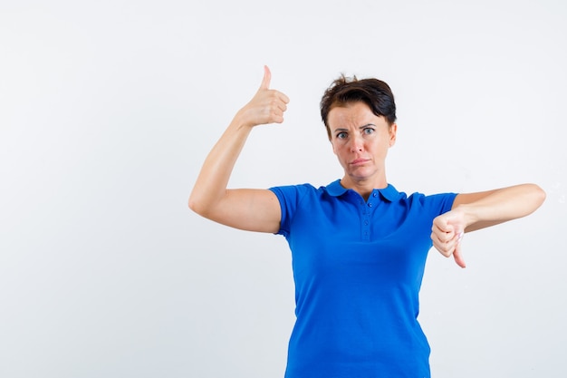 Mature woman showing thumbs up and down in blue t-shirt and looking hesitant , front view.