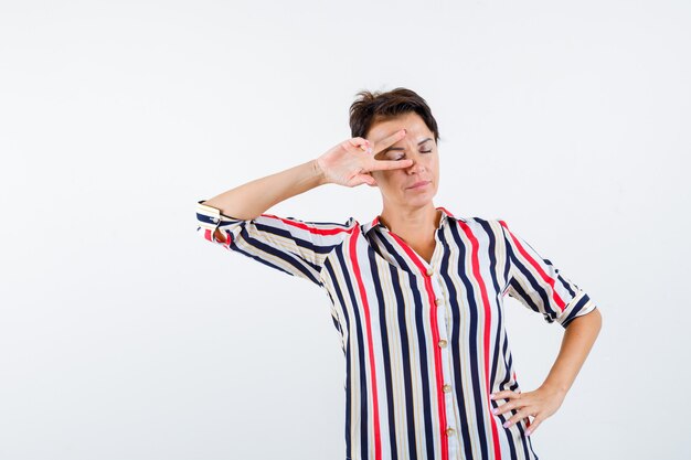 Mature woman showing v sign on eye while closing eyes, holding hand on waist in striped blouse and looking calm. front view.