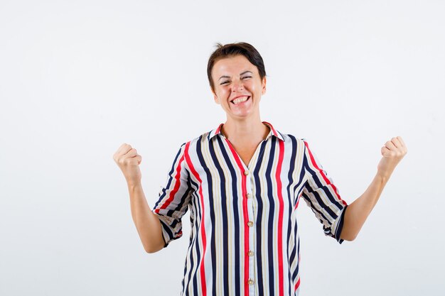 Mature woman showing winner gesture in striped blouse and looking lucky , front view.