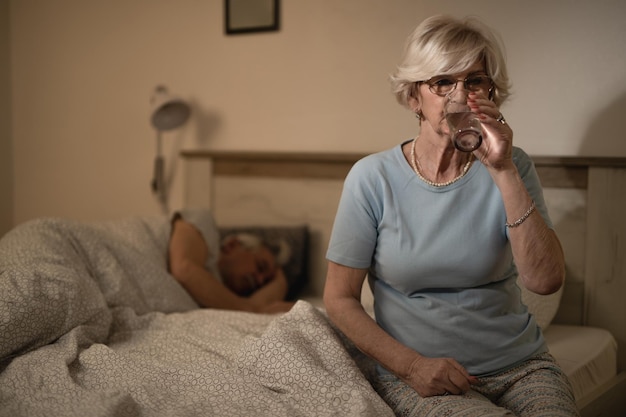 Foto gratuita donna matura seduta sul letto e bevendo un bicchiere d'acqua mentre suo marito dorme sullo sfondo