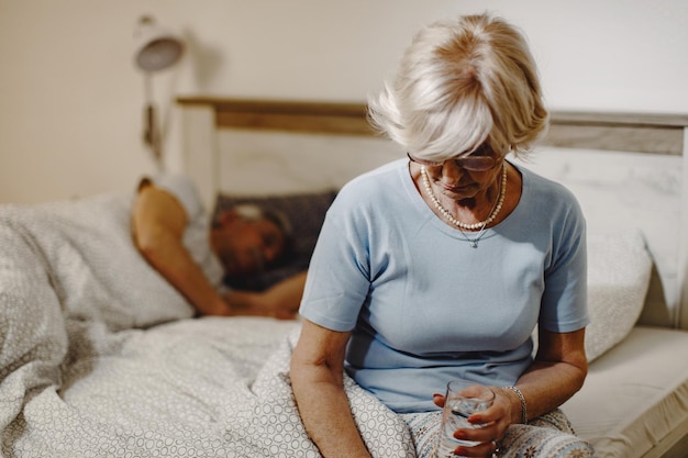 Бесплатное фото Зрелая женщина сидит на кровати и держит стакан воды. ее муж спит на заднем плане.