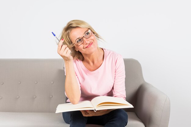 Mature woman sitting on sofa holding pen and an open book daydreaming