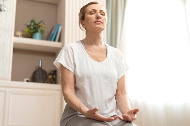 自宅で瞑想するスポーツウェアの成熟した女性。ヨガの練習。ヨガマットに目を閉じて座っています。ヨガのコンセプト。