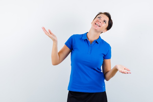 Mature woman spreading palms aside while looking up in blue t-shirt and looking hopeful , front view.