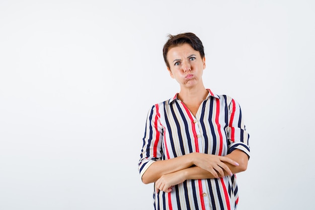 Mature woman standing arms crossed, puffing cheeks in striped blouse and looking annoyed. front view.