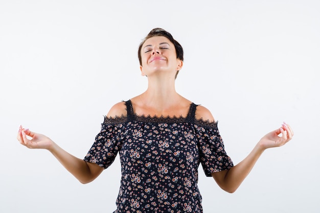 Free photo mature woman standing in meditating pose, closing eyes, smiling in floral blouse, black skirt and looking relaxed , front view.