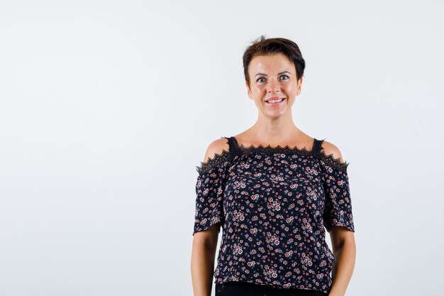 Mature woman standing straight and posing at camera in floral blouse, black skirt and looking cheery. front view.