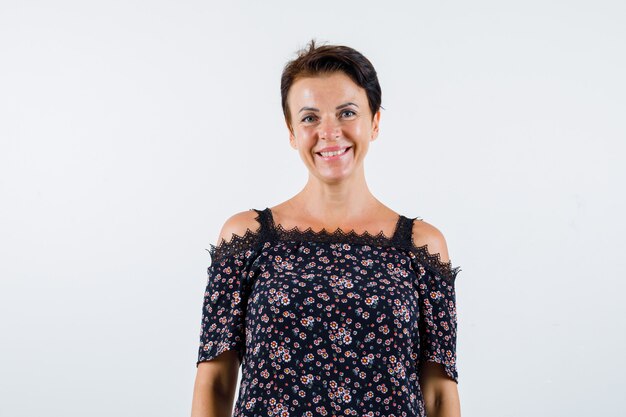 Mature woman standing straight and smiling in floral blouse, black skirt and looking confident. front view.