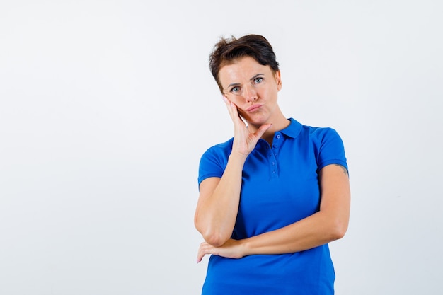 Mature woman standing in thinking pose in blue t-shirt and looking doubtful , front view.