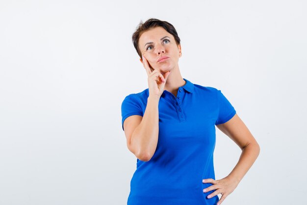 Mature woman standing in thinking pose in blue t-shirt and looking doubtful , front view.