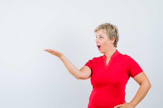 Зрелая женщина протягивает руку в озадаченном жесте в красной футболке
