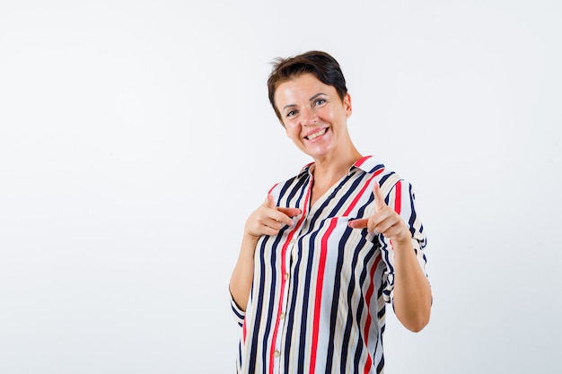 Зрелая женщина в полосатой блузке, указывая на камеру указательными пальцами и выглядя жизнерадостной, вид спереди.