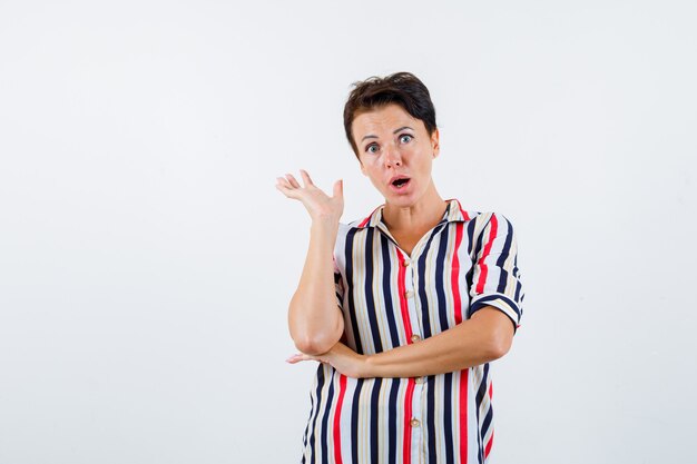 Mature woman in striped blouse raising one hand, holding another hand under elbow and looking surprised , front view.