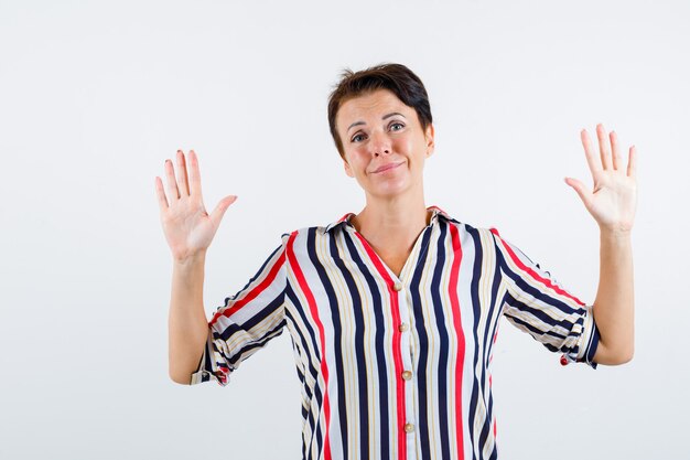 Mature woman in striped blouse raising palms in surrender gesture and looking happy , front view.