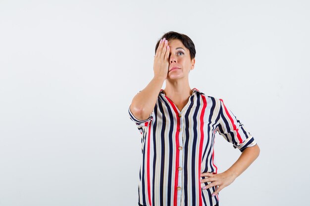 Mature woman in striped shirt covering eye with hand, holding hand on waist and looking serious , front view.