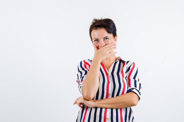 Mature woman in striped shirt covering mouth with hand, holding on hand under elbow and looking happy , front view.