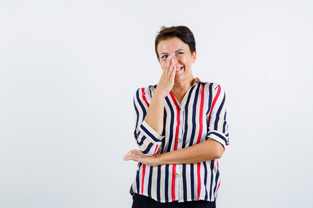 Mature woman in striped shirt covering mouth with hand, holding one hand under elbow and looking cheery , front view.