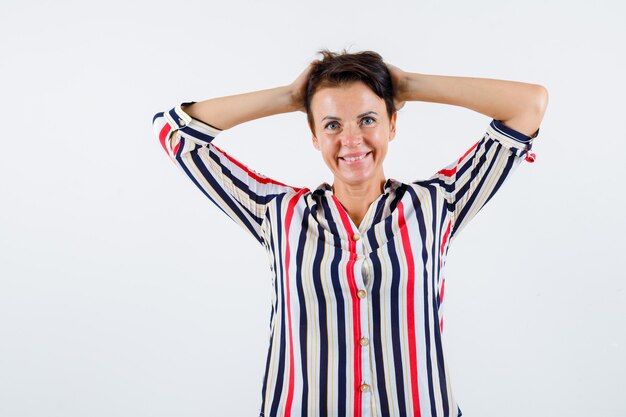 Donna matura in camicia a righe tenendo le mani sulla testa e guardando felice, vista frontale.