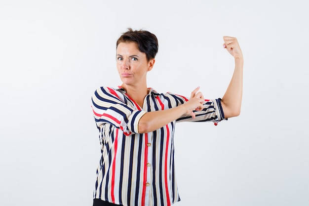 Зрелая женщина в полосатой рубашке показывает жест власти, измеряет размер бицепса и выглядит уверенно, вид спереди.