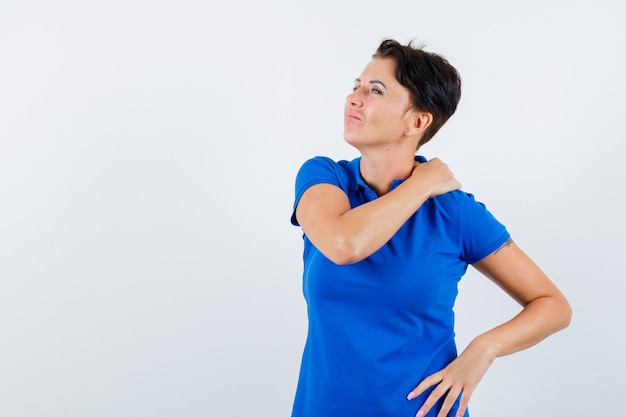 青いTシャツの肩の痛みに苦しんで疲れているように見える成熟した女性。正面図。