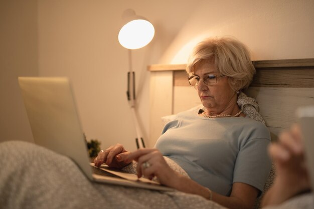 mature woman using laptop while lying down in bed in the evening