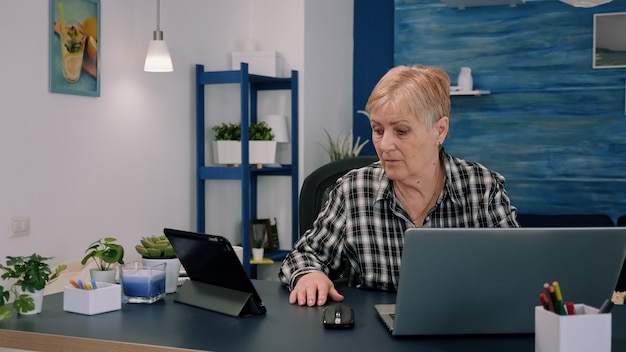 Зрелая женщина с помощью планшета и ноутбука одновременно анализирует финансовые графики, работая дома, сидя на рабочем месте