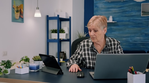 Mature woman using tablet and laptop in same time analysing financial graphs working from home sitting in workplace