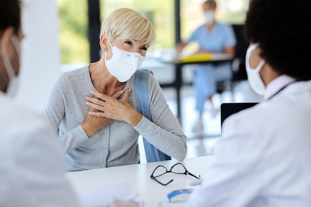 フェイスマスクをした成熟した女性がクリニックで医師と話しているときに胸の痛みを訴える